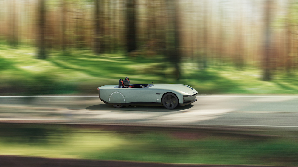 Image shows the design for an all-electric concept car called AURA, the sage-green, two-seater car is shown travelling along a road through a forest.