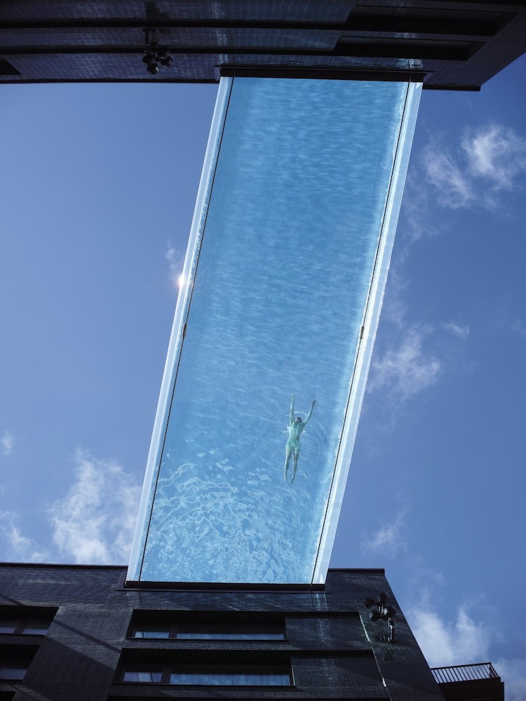 Image taken from underneath a 'sky pool'. This is a swimming pool with a transparent base layer that extends from one building to another at height. In the image we can see a single swimmer from below.